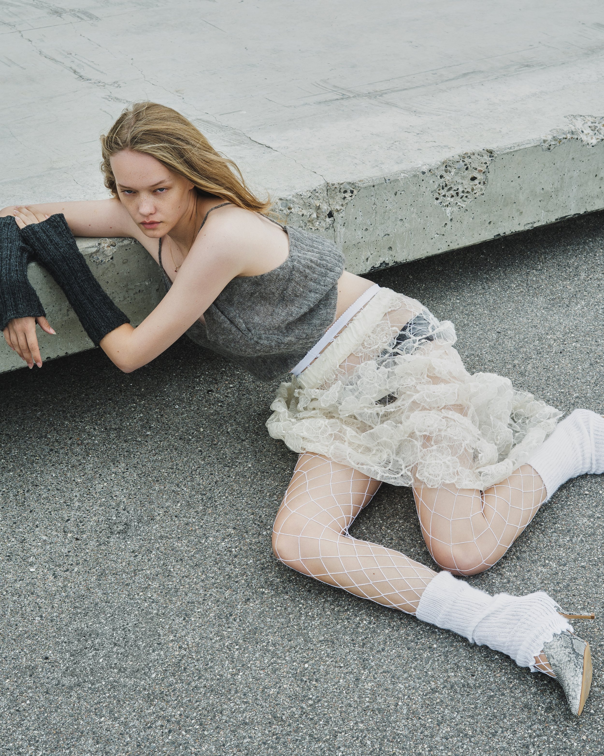 Woman with long blonde hair lying on the ground near a concrete ledge, wearing a gray tank top, lace skirt, fishnet stockings, and white socks with pointed shoes, in an outdoor urban setting.