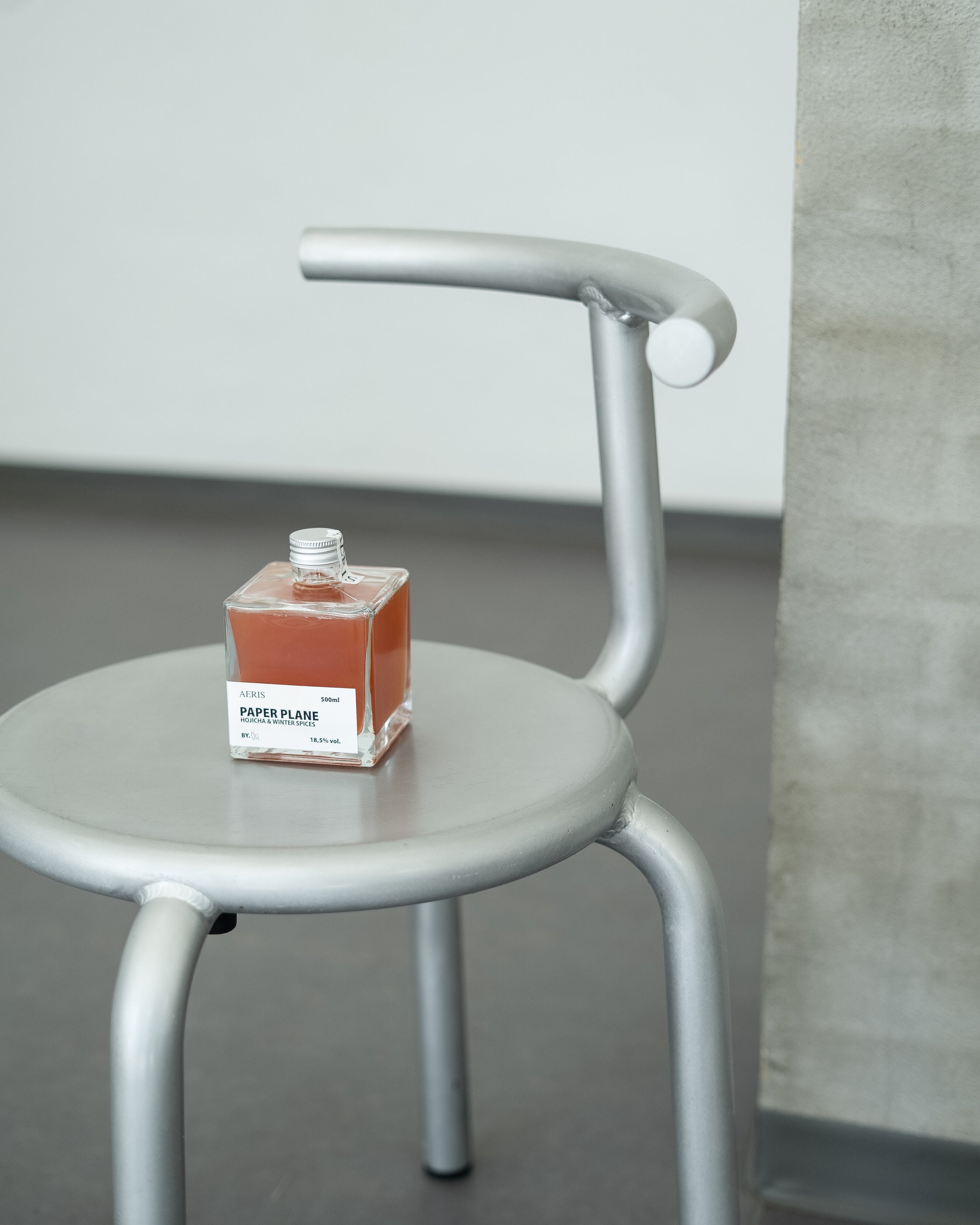 A silver metal faucet on a gray table, with a small transparent square bottle containing orange liquid labeled 'Paper Plane' perfume.