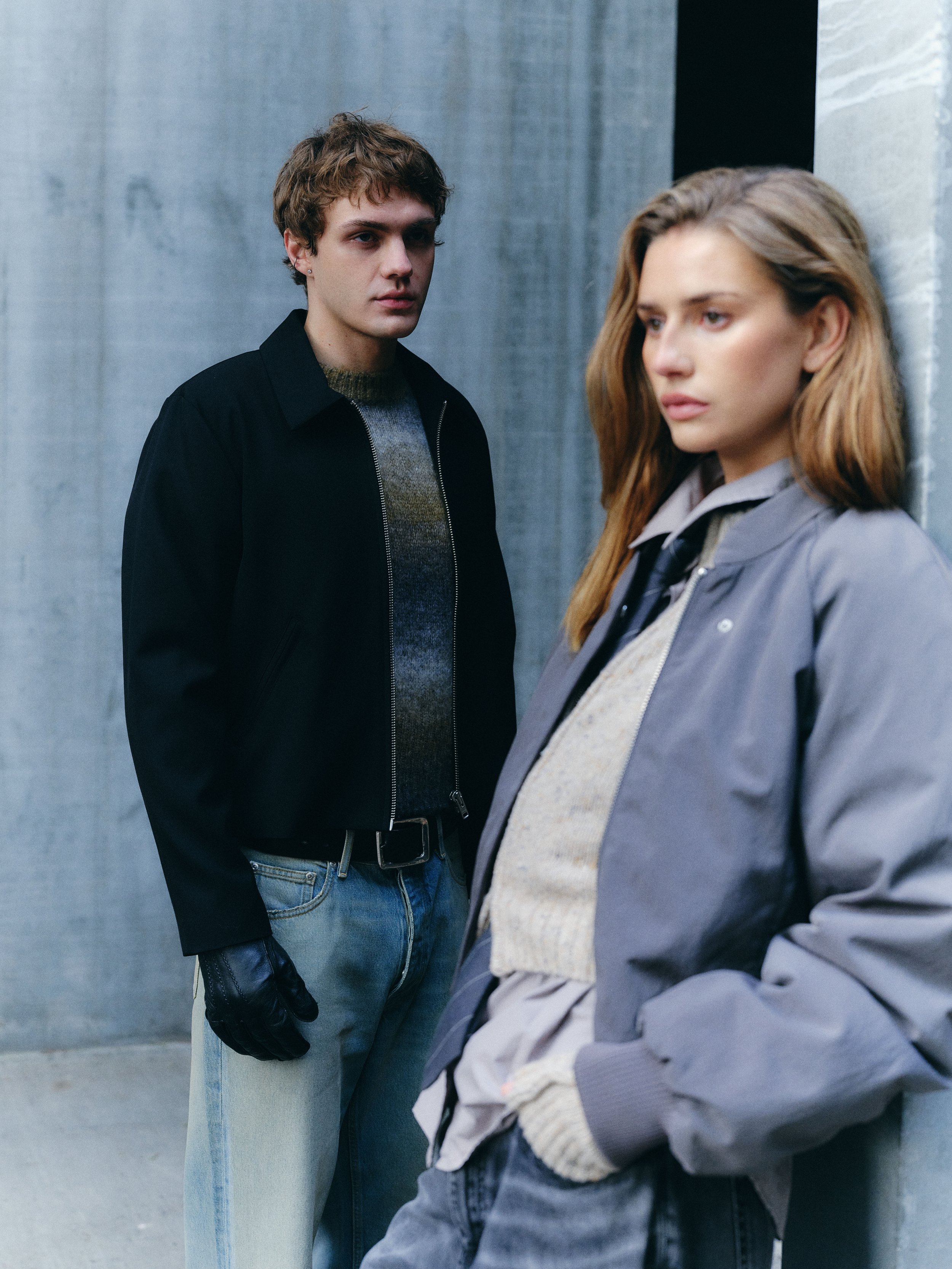 A young man and woman standing against a textured gray wall. The man is wearing a black jacket, gray sweater, and light blue jeans, with one hand in his pocket and a neutral expression. The woman is wearing a gray jacket, cream sweater, and dark jeans, leaning against the wall with her eyes looking off to the side.